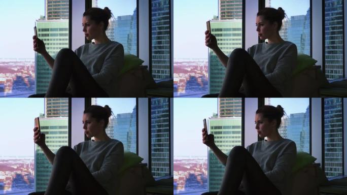 美丽的女性坐在窗台上，用智能手机拍照。女性正在使用小工具拍摄摩天大楼的照片