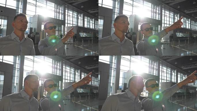 戴着墨镜的商人在终点站指着他的同事。两个年轻的商人在机场看航班时刻表。明亮的阳光透过背景的窗户照射。