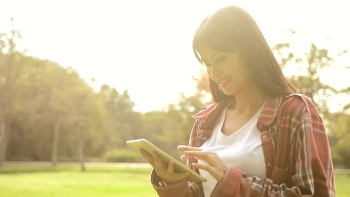 年轻漂亮的女人在公园的阳光早晨使用数字平板电脑，迷人的女人触摸屏数字平板电脑搜索信息