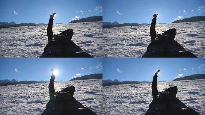 在阳光明媚的日子里，无法辨认的人躺在雪地上，用手指看着灿烂的阳光。年轻人在蓝天背景上触摸阳光的手。享