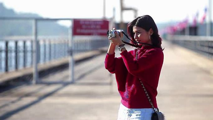 穿着红色连衣裙的年轻亚洲女孩在大坝上拿着相机拍照