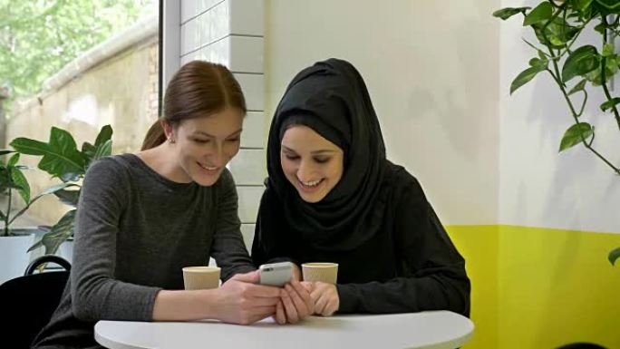 两个年轻的漂亮女人坐在咖啡馆里，其中一个是戴着头巾的穆斯林妇女，看着电话，笑着，惊讶地