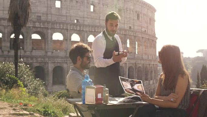 快乐的年轻夫妇游客在日落时坐在罗马罗马斗兽场前的酒吧餐厅向优雅的服务员订购食物和饮料