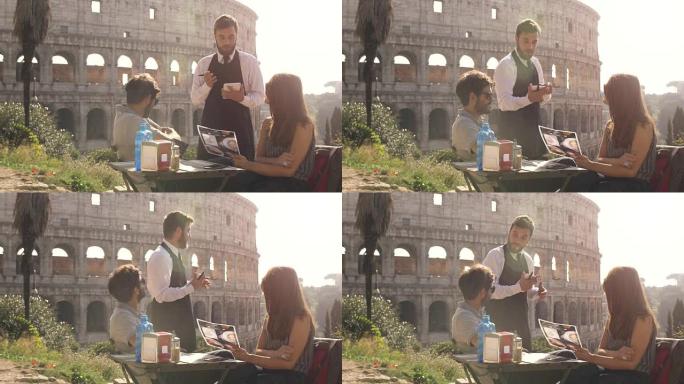 快乐的年轻夫妇游客在日落时坐在罗马罗马斗兽场前的酒吧餐厅向优雅的服务员订购食物和饮料