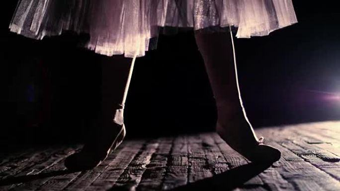 在聚光灯下，在旧剧院大厅的舞台上关闭。穿着白色芭蕾舞裙的芭蕾舞女演员，脚尖鞋抬起脚趾，优雅地进行某种