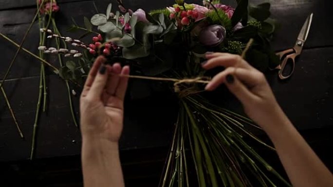 由花店装饰的一束美丽的鲜花躺在桌子上。女孩用绳子把它紧紧地系在茎上。从上方观看