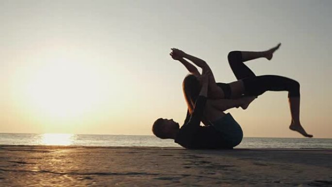 Acroyoga概念。两个运动的人在自然日出背景上成对练习瑜伽。美丽的年轻夫妇在海边做伸展运动。慢动