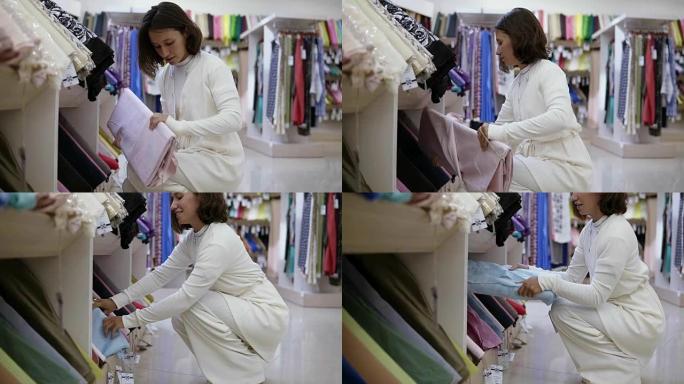 纺织店里华丽的年轻女子，背景上有各种纸巾。顾客决定选择哪种布料。考虑不同的类型。拿蓝色一卷。侧视图