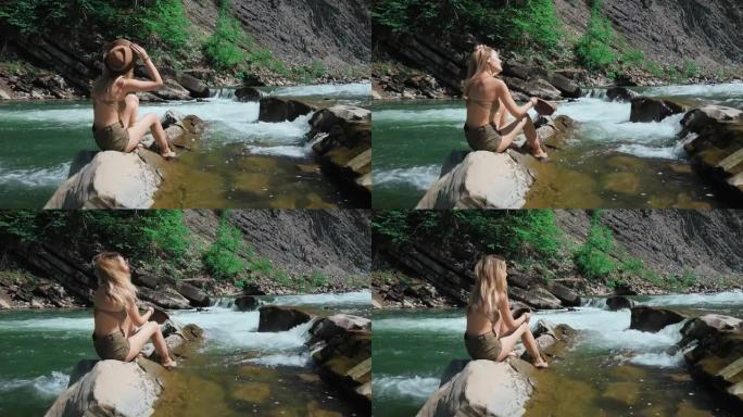 一名年轻女子坐在瑞士的一条山河岸边。脱下帽子，长出头发。自由和纯洁的概念，快乐。卸掉枷锁。慢动作