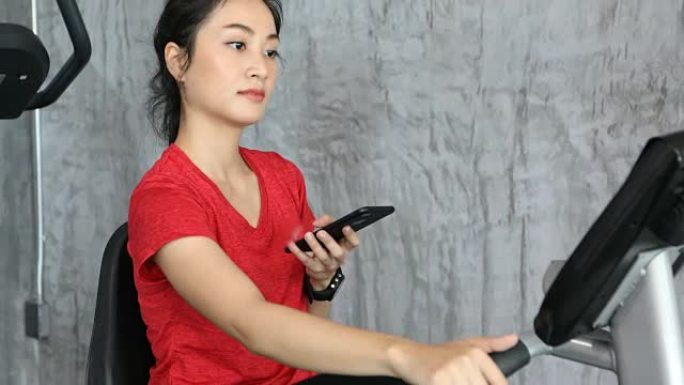 美女骑健身车骑自行车，在健身房使用智能手机