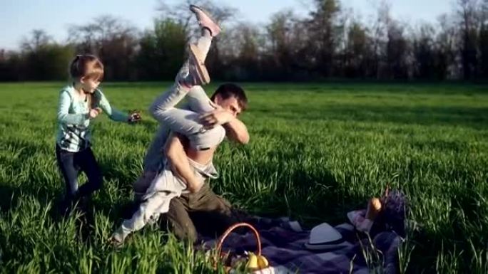 一位年轻的父母和他成年的女儿在草地上玩耍。他把年长的扔在肩上。年轻的一个嬉戏地跳上爸爸。慢动作