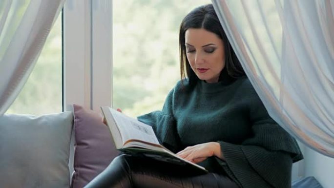 穿着绿色服装的美丽迷人的女人在舒适的窗台上放松，看书。微笑的女士。慢动作