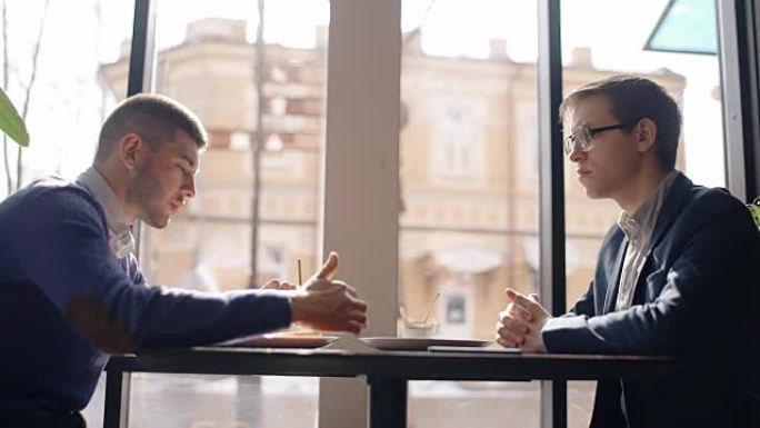 两个年轻人正在讨论创业，坐在咖啡馆的桌子旁，商人分享想法，交谈，在明亮的房间里通过窗户谈论新项目