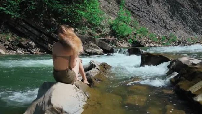 年轻女子坐在瑞士山河岸边的岩石上。脱下帽子，张开长发。自由和纯洁的概念，快乐。卸掉枷锁。慢动作