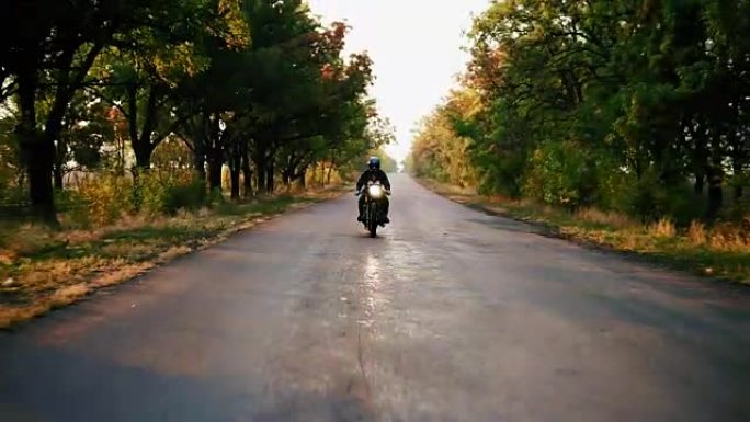 在秋天阳光明媚的日子里，穿着黑色头盔和皮夹克的时尚男人在柏油路上骑摩托车的前视图。美丽的树木，周围有