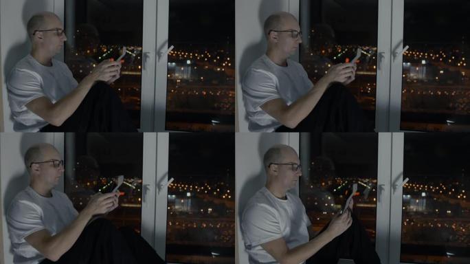 戴着眼镜的秃头男人看着平板电脑坐在窗台上，夜景
