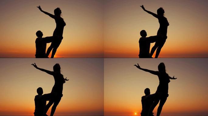 日落时在海边做双人瑜伽的年轻男女剪影。冥想。夫妇练习acro瑜伽。自然背景下的灵活性锻炼。慢动作