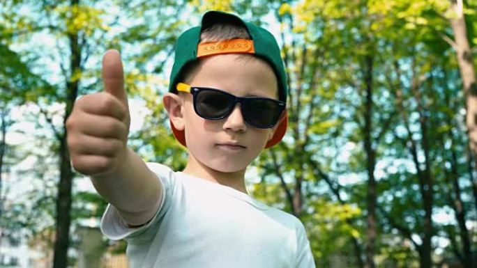一个戴着墨镜的酷男孩展示了一个班级，他的手指在绿色的树木背景下站在夏季公园里