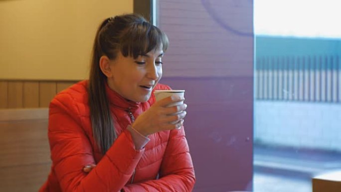 迷人的女人在咖啡馆喝咖啡。穿着红色泡泡夹克的高加索女性肖像，享受着咖啡香味，在窗户附近啜饮卡布奇诺咖