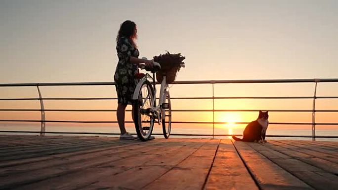 年轻迷人的女人穿着复古自行车和淡紫色花束，在日出或日落时靠近海边。女孩钦佩黎明，享受着在海洋海岸上生