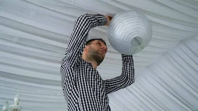 男子将彩色白色粉色灰色纸灯笼挂在天花板上。