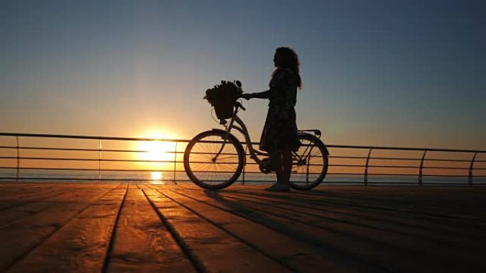 年轻迷人的女人穿着复古自行车的剪影，在日出或日落时在海边的木堤上行走。浪漫旅行理念。4k