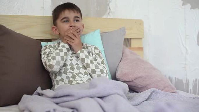 一个生病的小男孩坐在床上咳嗽，用毯子盖住自己。高清