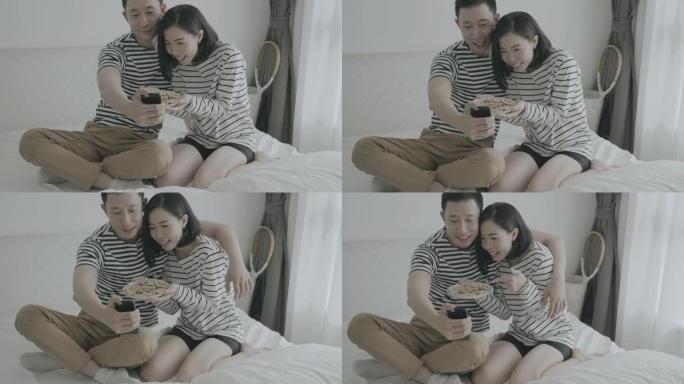 年轻夫妇在家中使用智能手机时吃零食，在社交网络中与朋友吃饭，交谈和展示时享受爱情和婚姻-社交媒体概念