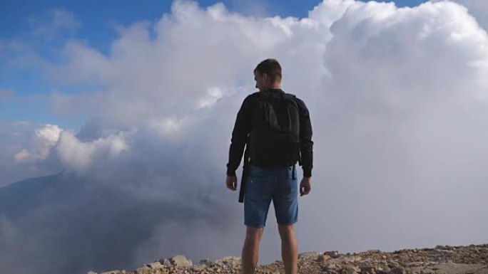 面目全非的年轻男性徒步旅行者，背包站在山的边缘，享受美丽的风景。背景多云的天空。旅游概念。后视图慢动