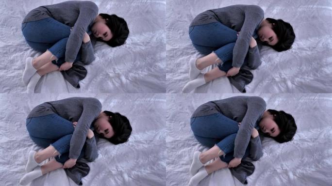 顶拍年轻悲伤的亚洲女孩躺在床上，用胳膊抱着膝盖，不开心，白色卧室50 fps