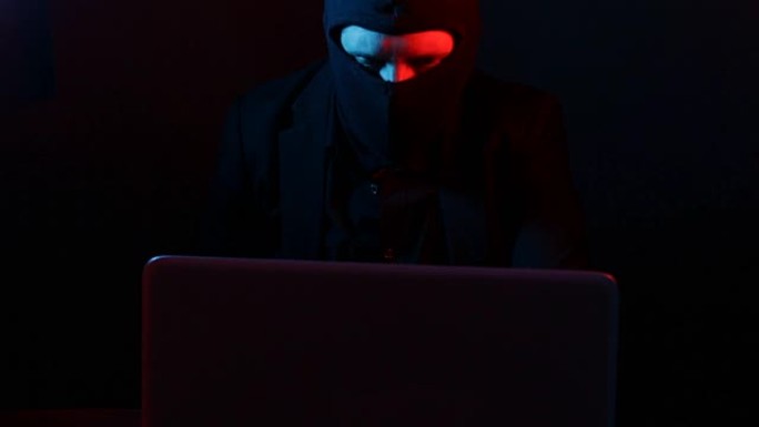 穿着西装的愤怒的计算机黑客从红色和蓝色灯光照亮的笔记本电脑中窃取数据