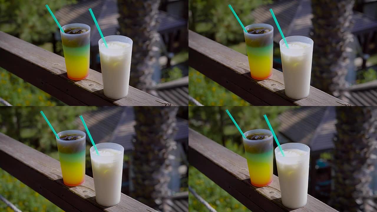 在阳光明媚的夏天，一杯多汁的分层鸡尾酒和一杯乳白色混合饮料站在阳台上的扶手上