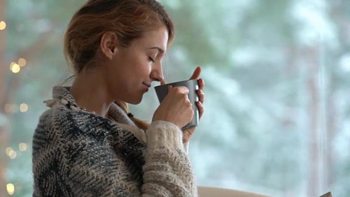 舒适的冬季生活方式。年轻快乐的女人喝杯咖啡穿着针织毛衣坐在家里的大窗户与冬天的雪背景