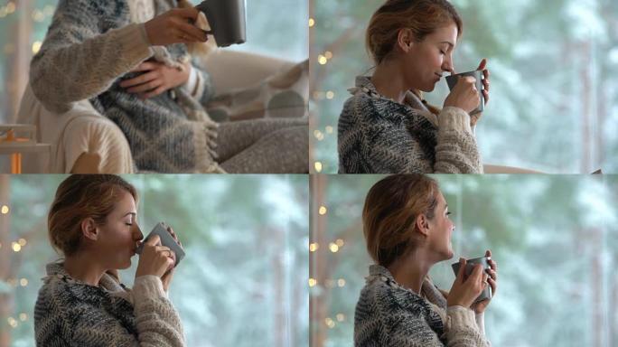 舒适的冬季生活方式。年轻快乐的女人喝杯咖啡穿着针织毛衣坐在家里的大窗户与冬天的雪背景