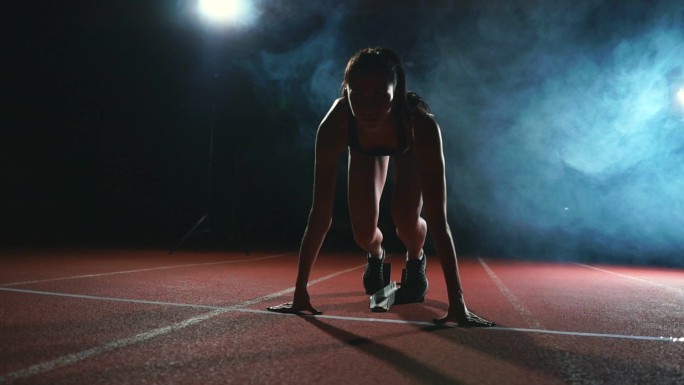 黑暗背景下的女运动员正准备从跑步机上的垫子上跑越野短跑