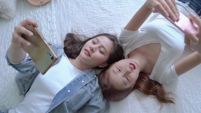 两个漂亮的亚洲朋友在床上玩手机的俯视视频，消费技术的概念正在迅速发展。