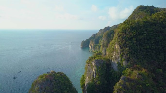 空中：壮观的夏日阳光照耀在热带绿地覆盖的大型石灰岩悬崖上，旅游观光船在美丽的平静海洋中航行。令人叹为