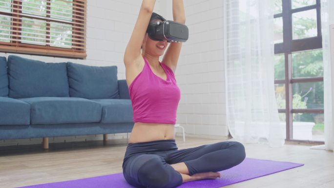 年轻的亚洲少女使用虚拟现实模拟器，同时在客厅练习瑜伽，女性锻炼健康在家里。生活方式女性锻炼未来技术理