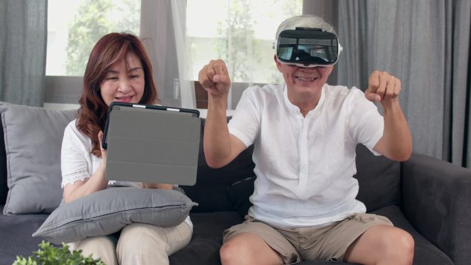 亚洲老年夫妇使用平板电脑和虚拟现实模拟器在客厅玩游戏，夫妻俩一起开心地躺在家里的病床上。生活方式 高