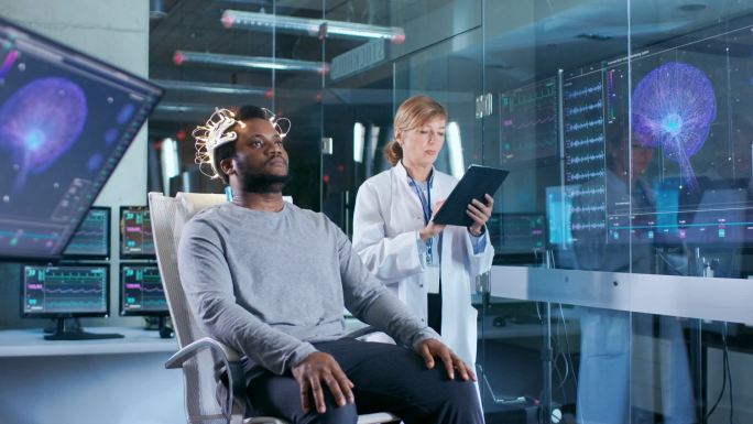 男子穿着脑波扫描耳机坐在椅子上, 而科学家与平板电脑监督过程。现代脑研究中的实验室监测显示英语阅读和