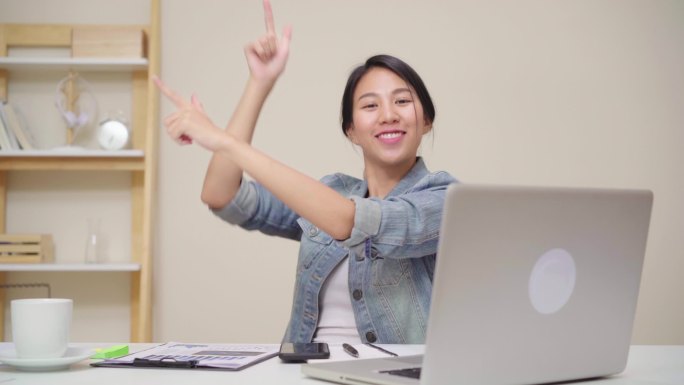 年轻的亚洲妇女在家里客厅的办公桌上使用笔记本电脑工作。亚洲女商人成功庆祝在家庭办公室快乐跳舞的感觉。