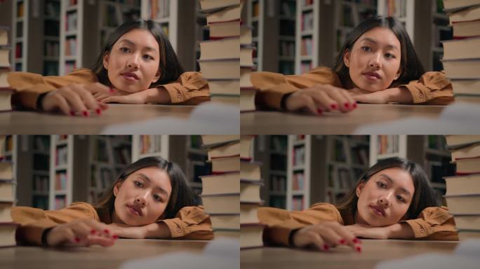 疲倦的沉思的年轻亚洲妇女躺在书桌上准备在大学图书馆准备考试的书无聊的体贴懒惰的学生女孩看着远处想着做