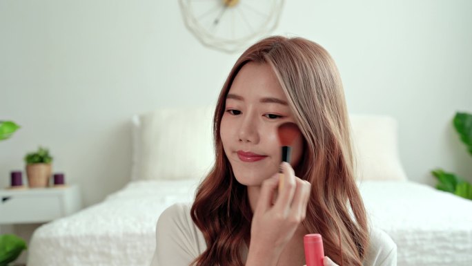  漂亮的女人坐在卧室里用化妆品刷把脸弄红了。亚洲女人自己化妆.
