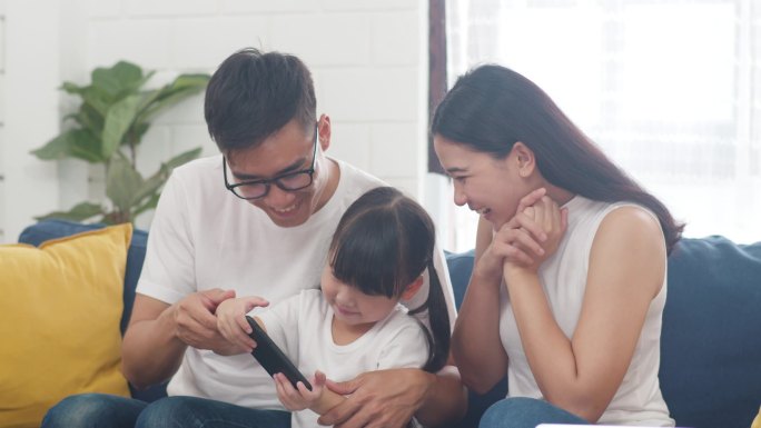 快乐的亚洲家庭爸爸、妈妈和女儿在网上玩着好玩的游戏、坐在家里的沙发上。自我隔离、待在家里、与他人保持