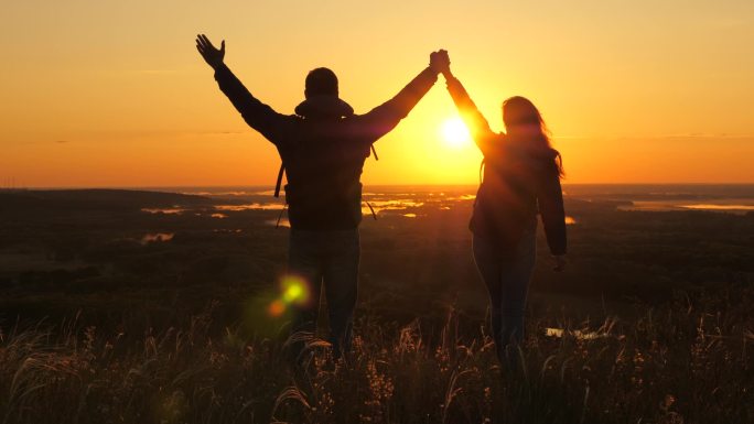 旅行者，一个男人和一个女人，背着背包，在黎明的光芒中走到山边，举起双手，享受胜利，美丽的阳光和风景。
