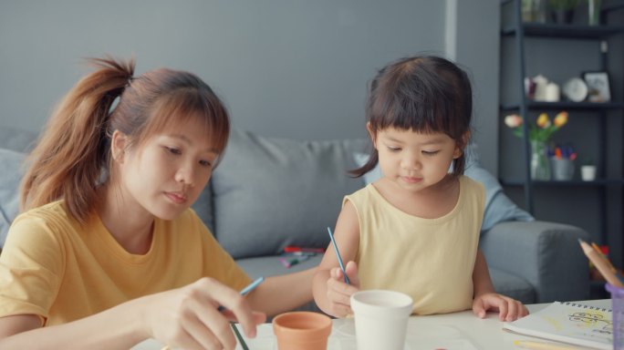 快乐快乐的亚洲家庭妈妈教蹒跚学步的小女孩画瓷壶在客厅的桌上玩乐放松。在一起度过的时间、社会距离、检疫