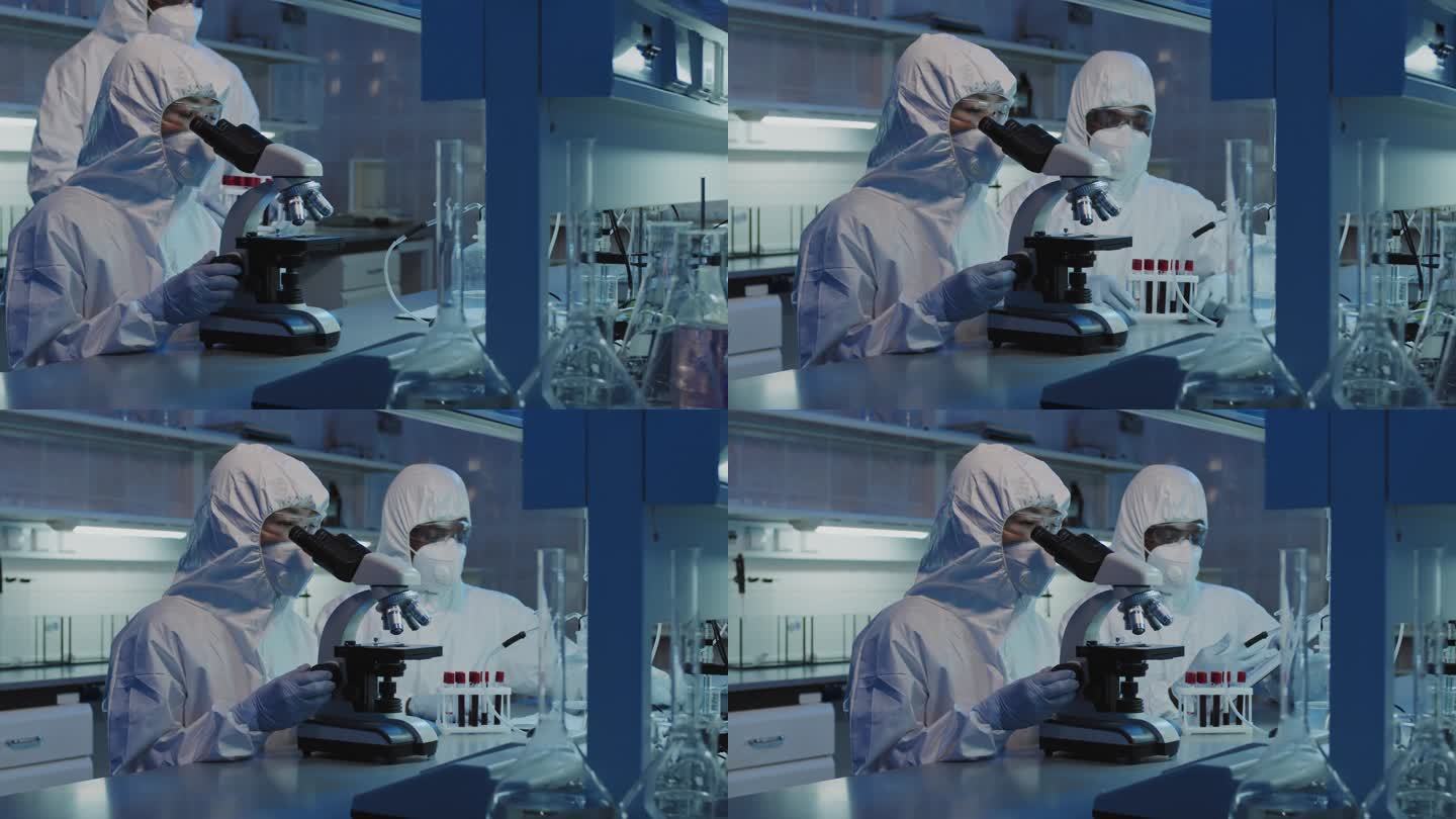 男同事在实验室的显微镜下、穿着防护服、护目镜、面罩和手套、拿着装有与她交谈的血液的试管架、对无法辨认