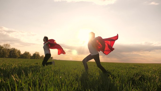 快乐的女孩扮演超级英雄，她们穿着红色斗篷在绿地上奔跑，风中飘扬着斗篷。孩子们的游戏和梦想。慢动作青少