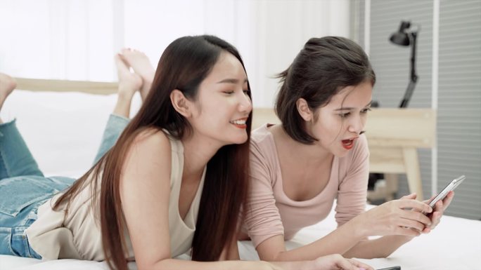 两个亚洲女孩躺在床上，在卧室里通过视频聊天与朋友聊天.