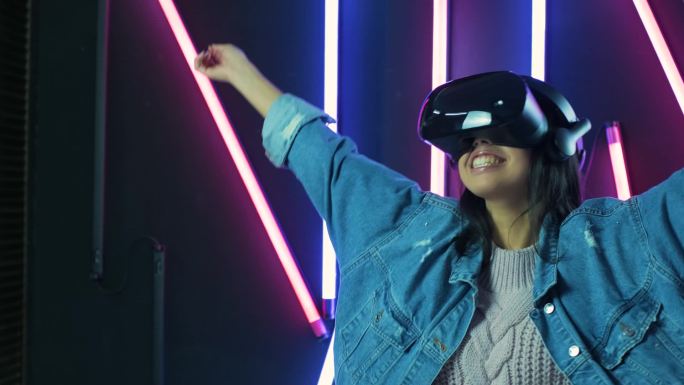 快乐的年轻女人环顾四周，想知道在VR耳机里有多神奇。彩灯背景下的虚拟现实头盔